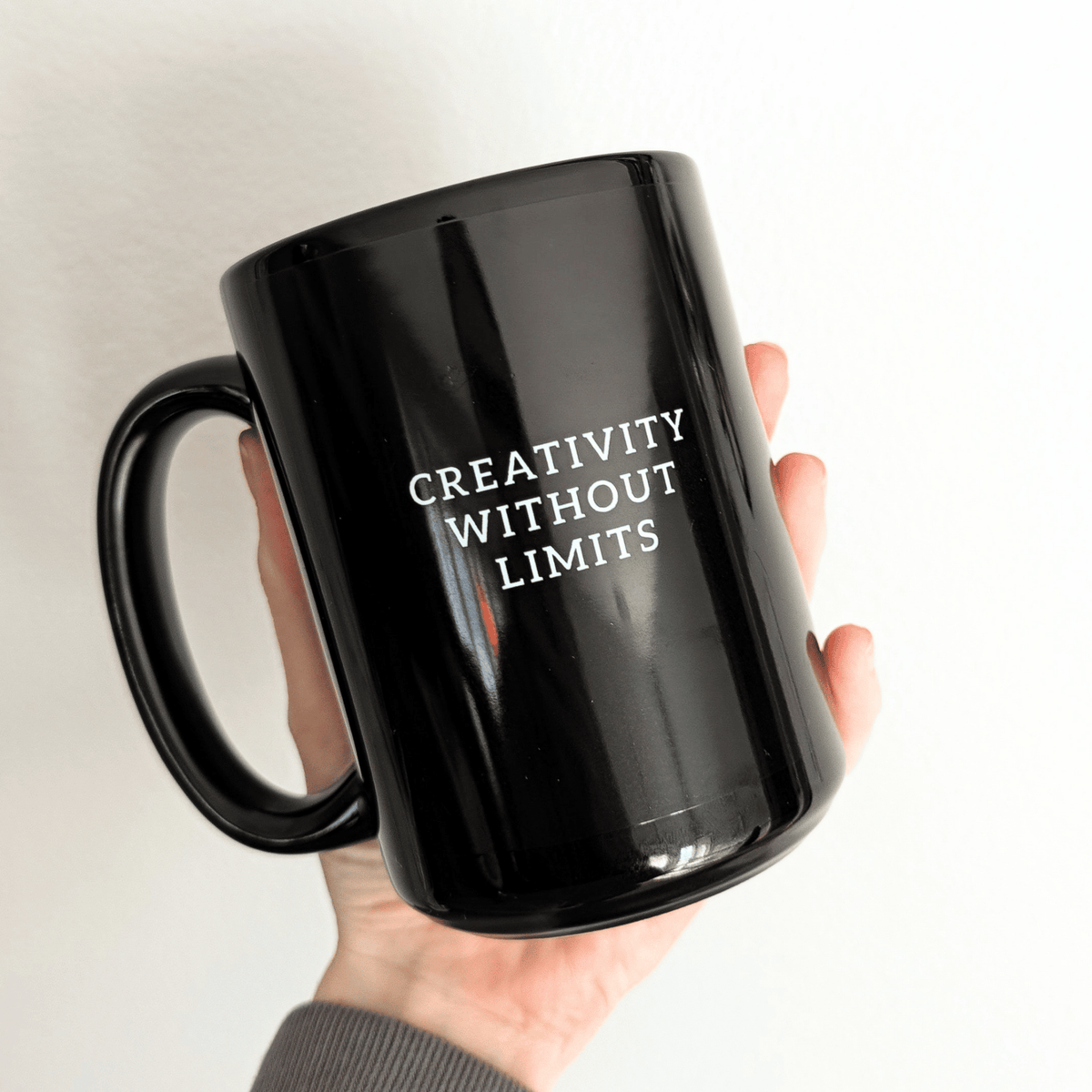 A hand holding a glossy black ceramic mug with the phrase 'CREATIVITY WITHOUT LIMITS' printed in white capital letters on the front, set against a solid white background