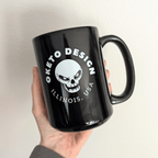 A hand holding a glossy black ceramic mug with a white printed graphic featuring a skull and the text 'OKETO DESIGN' and 'ILLINOIS, USA', shown on a white background