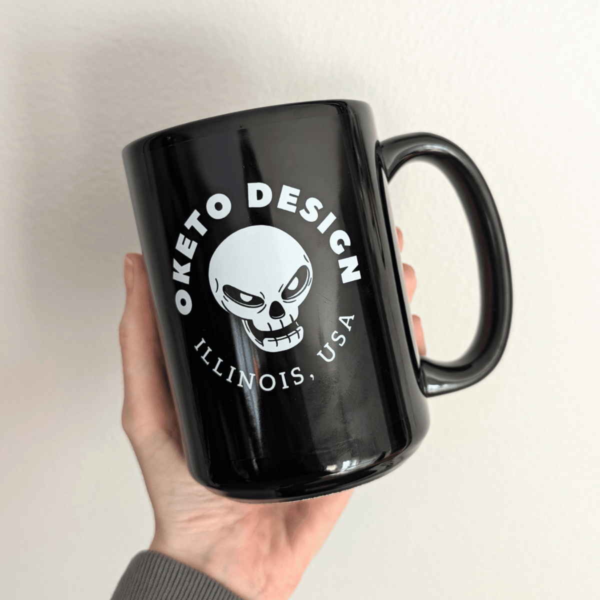 A hand holding a glossy black ceramic mug with a white printed graphic featuring a skull and the text 'OKETO DESIGN' and 'ILLINOIS, USA', shown on a white background