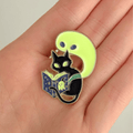 A hand holding a stylized enamel pin featuring a black cat reading a glowing book on a white background