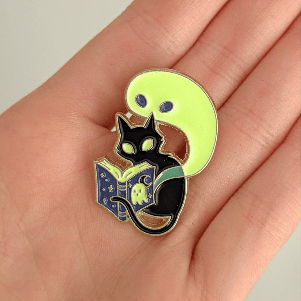 A hand holding a stylized enamel pin featuring a black cat reading a glowing book on a white background
