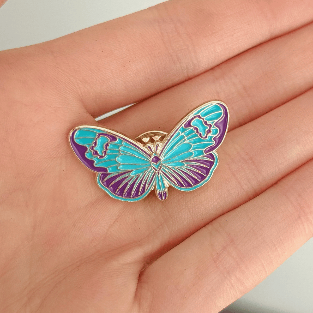 A hand holding a blue butterfly enamel pin with purple and gold details on a plain white background