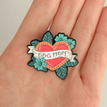 A hand holding a traditional tattoo-style enamel pin featuring a red heart with a gold border, teal flowers, and teal leaves on a white background