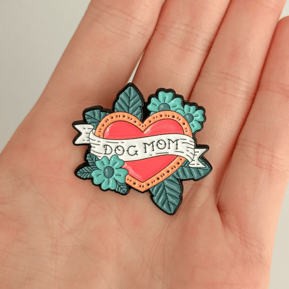A hand holding a traditional tattoo-style enamel pin featuring a red heart with a gold border, teal flowers, and teal leaves on a white background