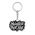 An acrylic keychain featuring the phrase 'Manifest That Shit' in white cursive lettering on a black background with a silver metal split ring and chain
