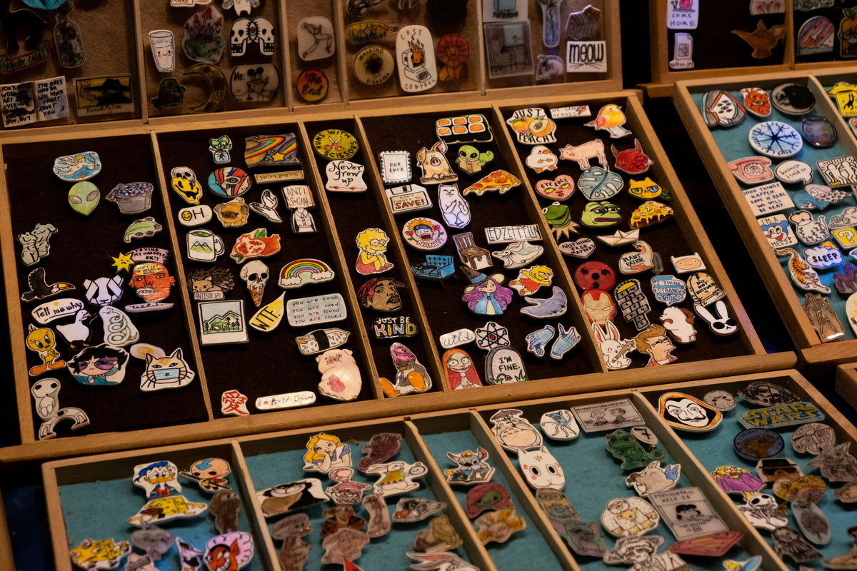 A display of numerous colorful enamel lapel pins arranged in wooden trays, featuring various cartoon, animal, and graphic designs