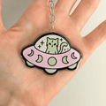 A hand holding a cute acrylic keychain featuring a cartoon green cat sitting inside a pink UFO with moon patterns, attached to a silver metal key ring on a plain white background
