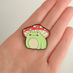 A hand holding a cute cartoon frog pin sitting under a red mushroom cap, shown on a solid white background