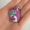 A hand holding a purple juice carton pin with a white goose head graphic, centered on a solid white background