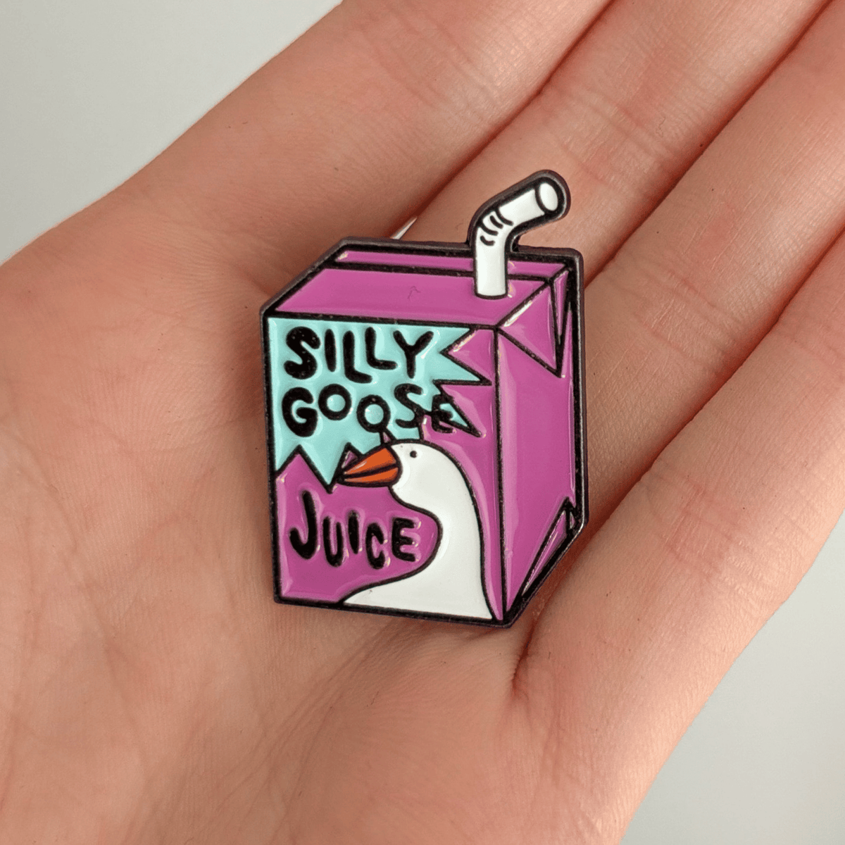 A hand holding a purple juice carton pin with a white goose head graphic, centered on a solid white background