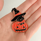 A hand holding a Halloween-themed enamel pin featuring a black cat wearing a witch hat and sitting on a carved orange pumpkin, shown on a solid white background