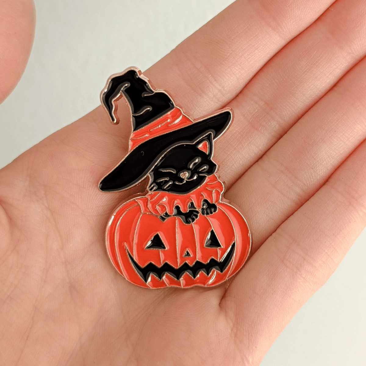 A hand holding a Halloween-themed enamel pin featuring a black cat wearing a witch hat and sitting on a carved orange pumpkin, shown on a solid white background