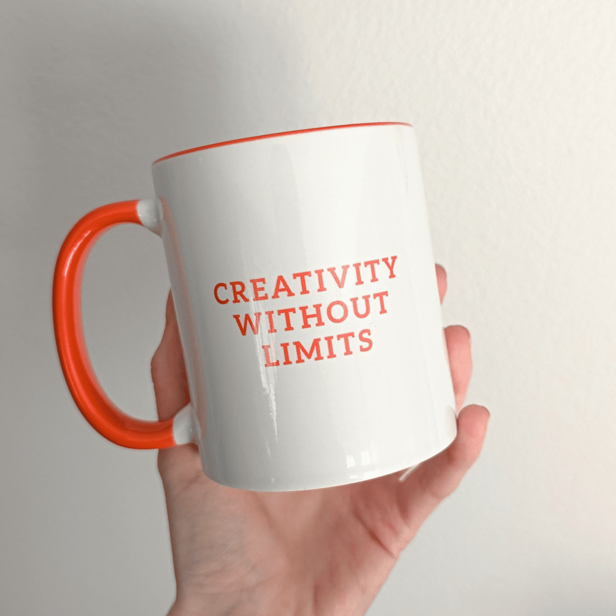 A hand holding a white mug with orange handle and text 'Creativity Without Limits' on a white background