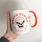 A hand holding a white ceramic mug with a bright orange handle and interior, featuring a skull graphic and the text 'OKETO DESIGN' and 'ILLINOIS, USA' in orange on the front