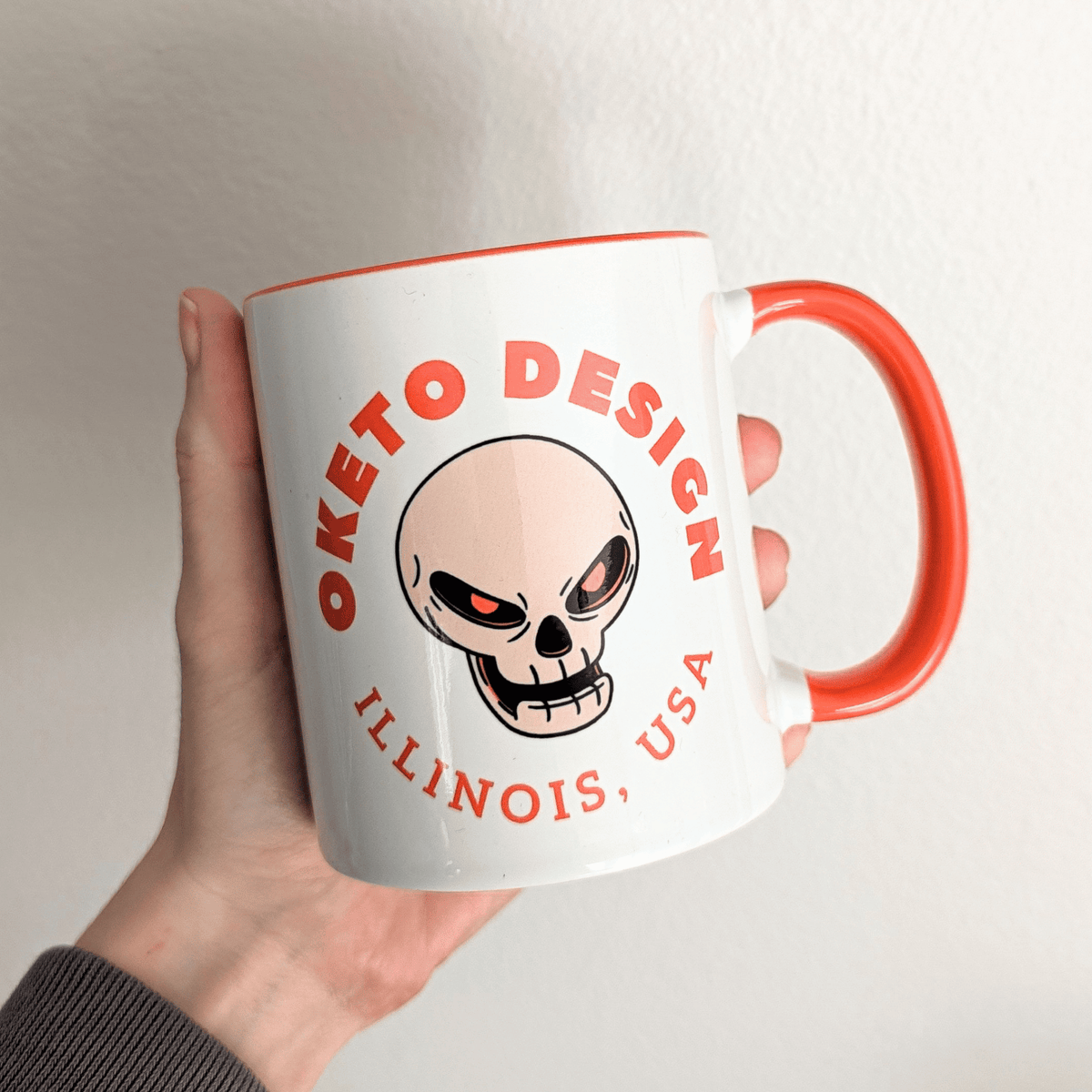A hand holding a white ceramic mug with a bright orange handle and interior, featuring a skull graphic and the text 'OKETO DESIGN' and 'ILLINOIS, USA' in orange on the front