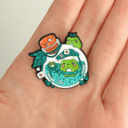 A hand holding a nature-themed enamel pin with frogs swimming inside a potion bottle on a white background