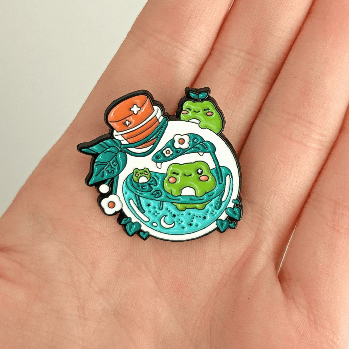 A hand holding a nature-themed enamel pin with frogs swimming inside a potion bottle on a white background