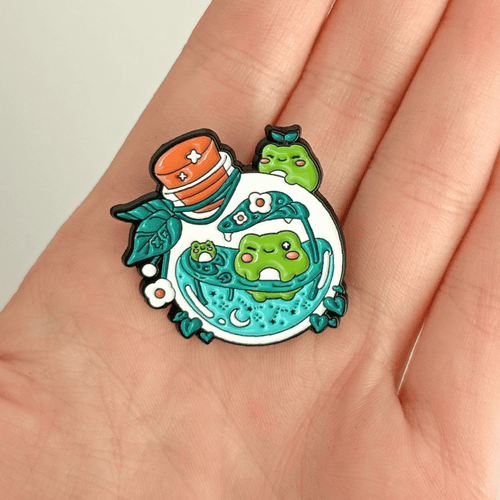 A hand holding a nature-themed enamel pin with frogs swimming inside a potion bottle on a white background