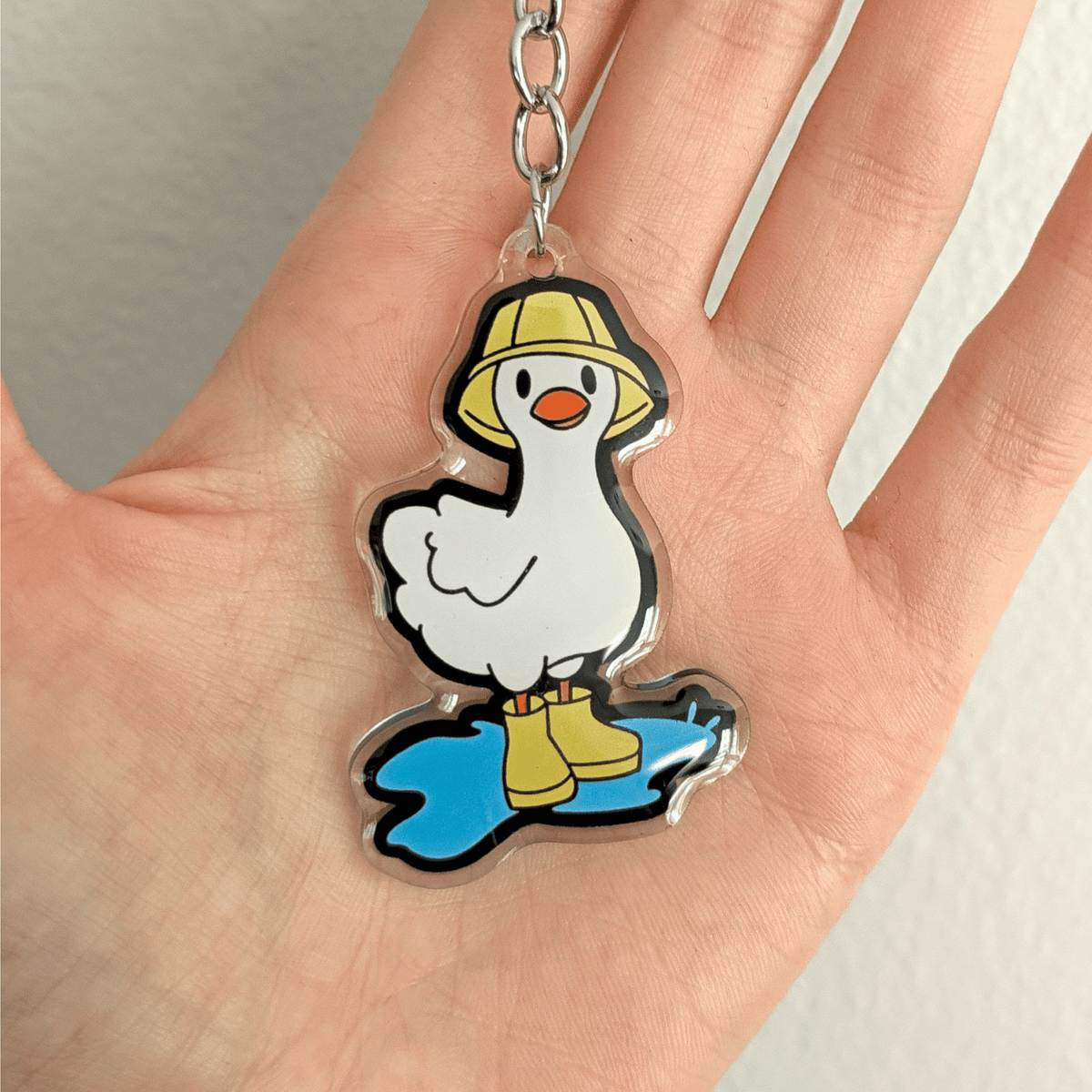 A hand holding a cartoon duck keychain charm with a white body, yellow bucket hat, yellow rain boots, and a blue puddle, attached to a silver metal key ring and chain on a plain white background