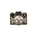A camera-shaped enamel pin featuring a black body with gold-tone outlines and naturalistic features set against a solid white background