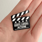 A hand holding a black clapperboard pin with the words 'LIGHTS, CAMERA ACTION!' on a white background