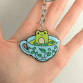 A hand holding a clear acrylic keychain charm shaped like a light blue teacup with green leaf patterns, featuring a cute green frog sitting in the center, attached to a chain and a split key ring on a white background
