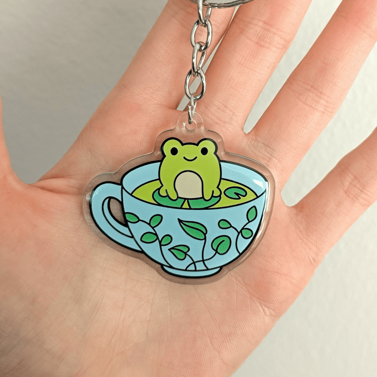 A hand holding a clear acrylic keychain charm shaped like a light blue teacup with green leaf patterns, featuring a cute green frog sitting in the center, attached to a chain and a split key ring on a white background