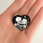 A hand holding a heart-shaped enamel pin featuring white skeletons embracing and kissing, centered on a solid white backdrop