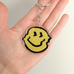 A hand holding a yellow smiley face charm keychain on a split ring clip with chain on a white background